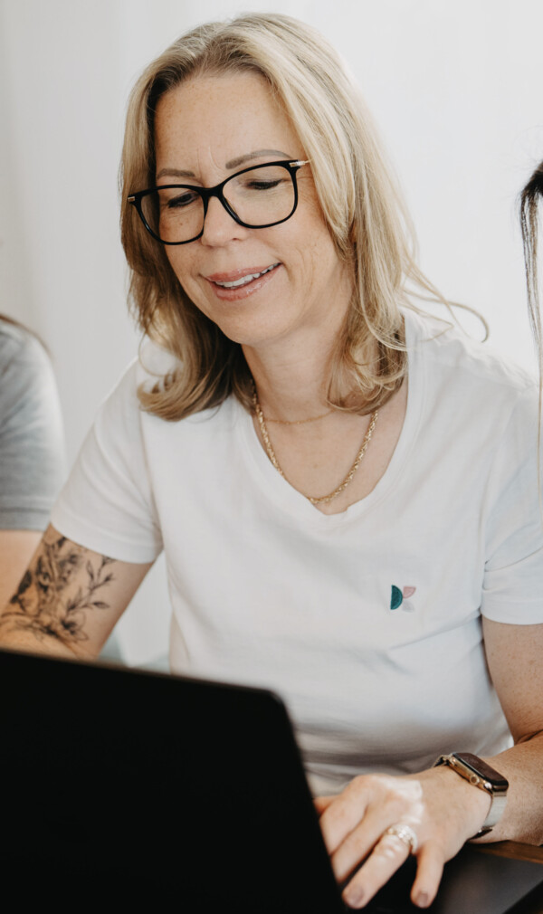 Drei Frauen in wei&szlig;en Shirts und grauem Poloshirt blicken konzentriert gemeinsam auf einen Laptop-Bildschirm.