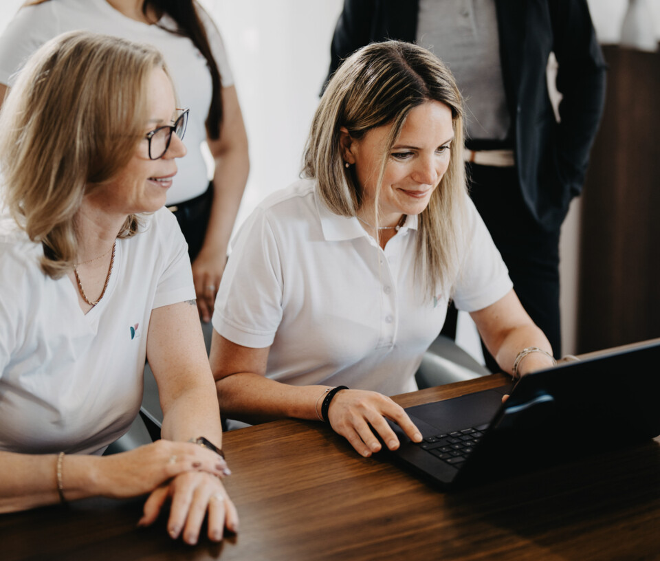 Andrea Hirschenberger und Tanja Breyer arbeiten l&auml;chelnd gemeinsam an einem Laptop an einem Holztisch.