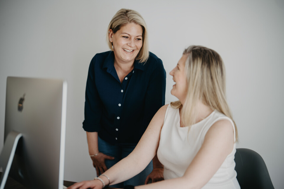 Charlene Antes und Melanie Siebert arbeiten gemeinsam am Computer. Charlene Antes im dunkelblauen Hemd beugt sich l&auml;chelnd zu ihrer Kollegin.