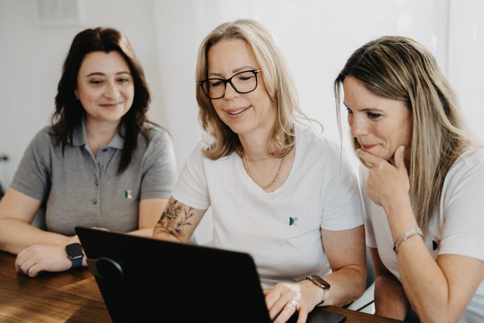 Drei Frauen in wei&szlig;en Shirts und grauem Poloshirt blicken konzentriert gemeinsam auf einen Laptop-Bildschirm.