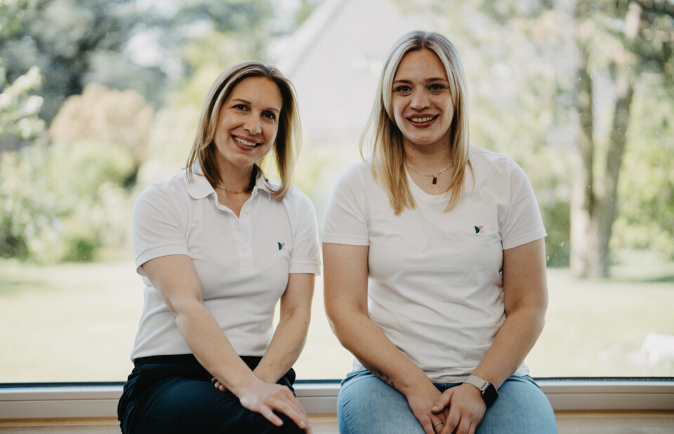 Christiane Berger und Jana Hellmann sitzen l&auml;chelnd vor einem hellen Fenster.