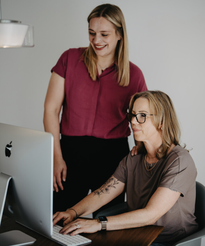 Andrea Hirschberger arbeitet am Computer, Jana Hellmann in pinker Bluse steht l&auml;chelnd hinter ihr.