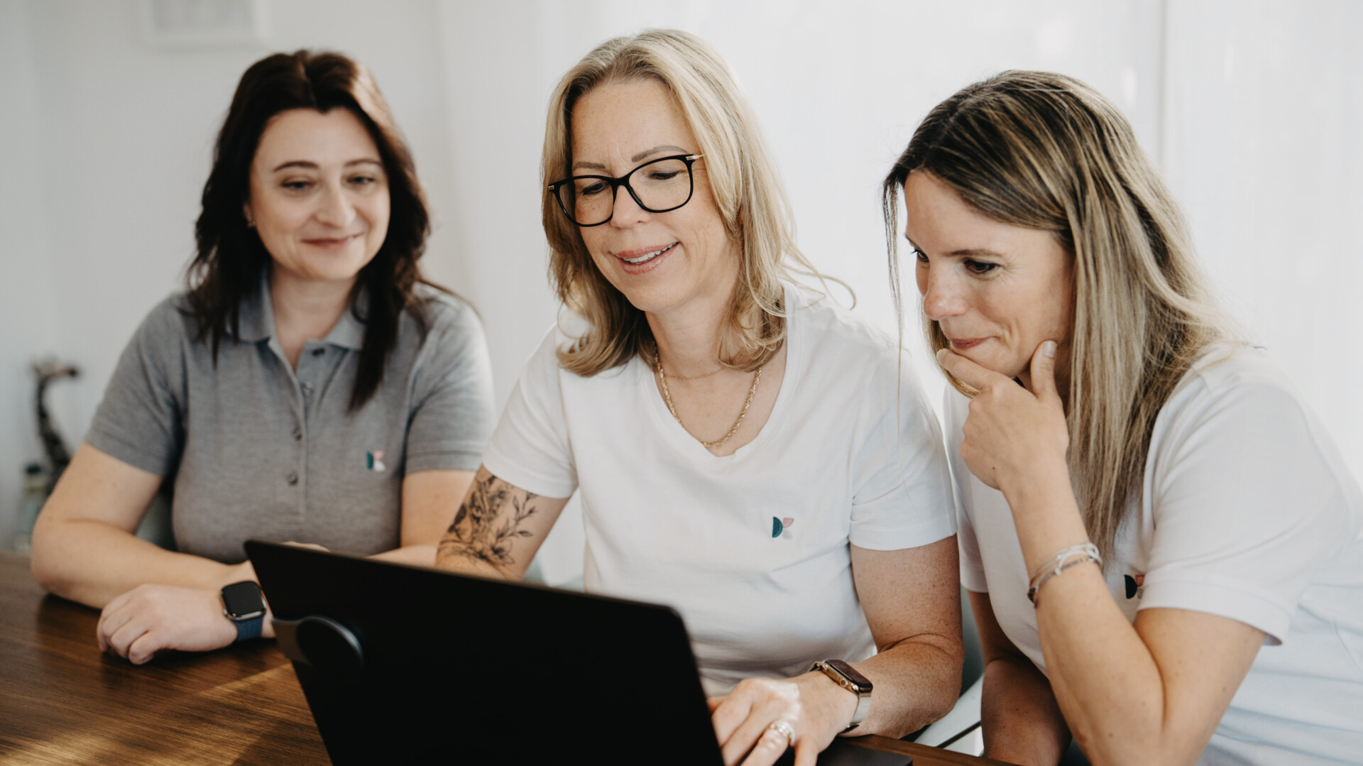 Drei Frauen in wei&szlig;en Shirts und grauem Poloshirt blicken konzentriert gemeinsam auf einen Laptop-Bildschirm.
