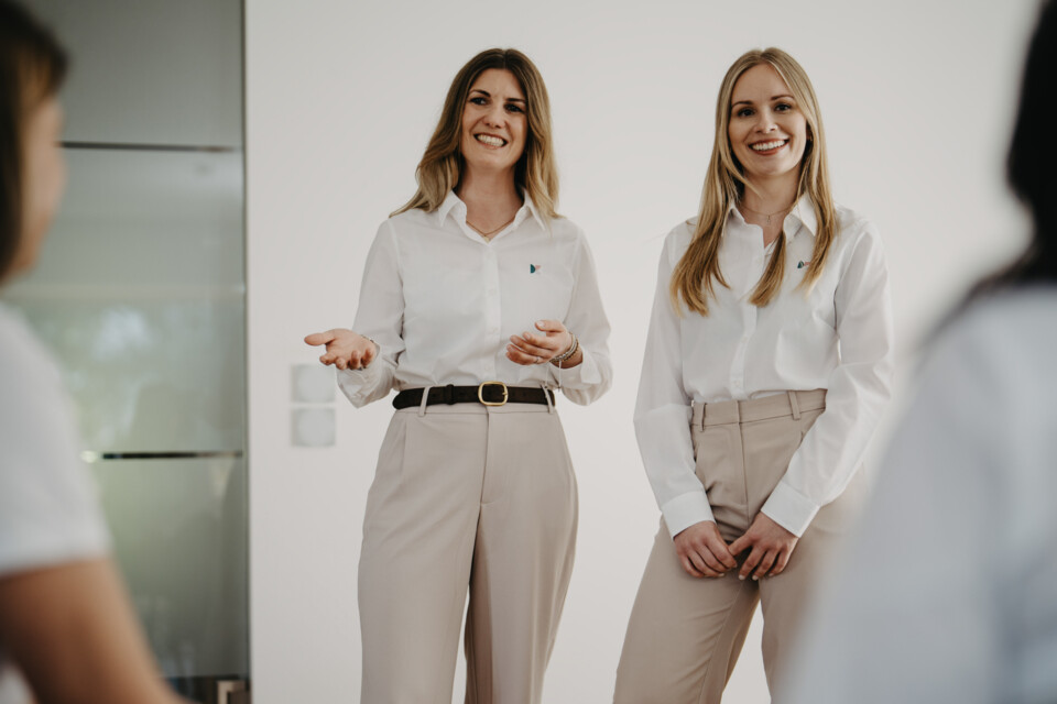 Zwei l&auml;chelnde Frauen in wei&szlig;en Blusen und beigen Hosen sprechen bei einer Pr&auml;sentation vor Kollegen in hellem B&uuml;ro.