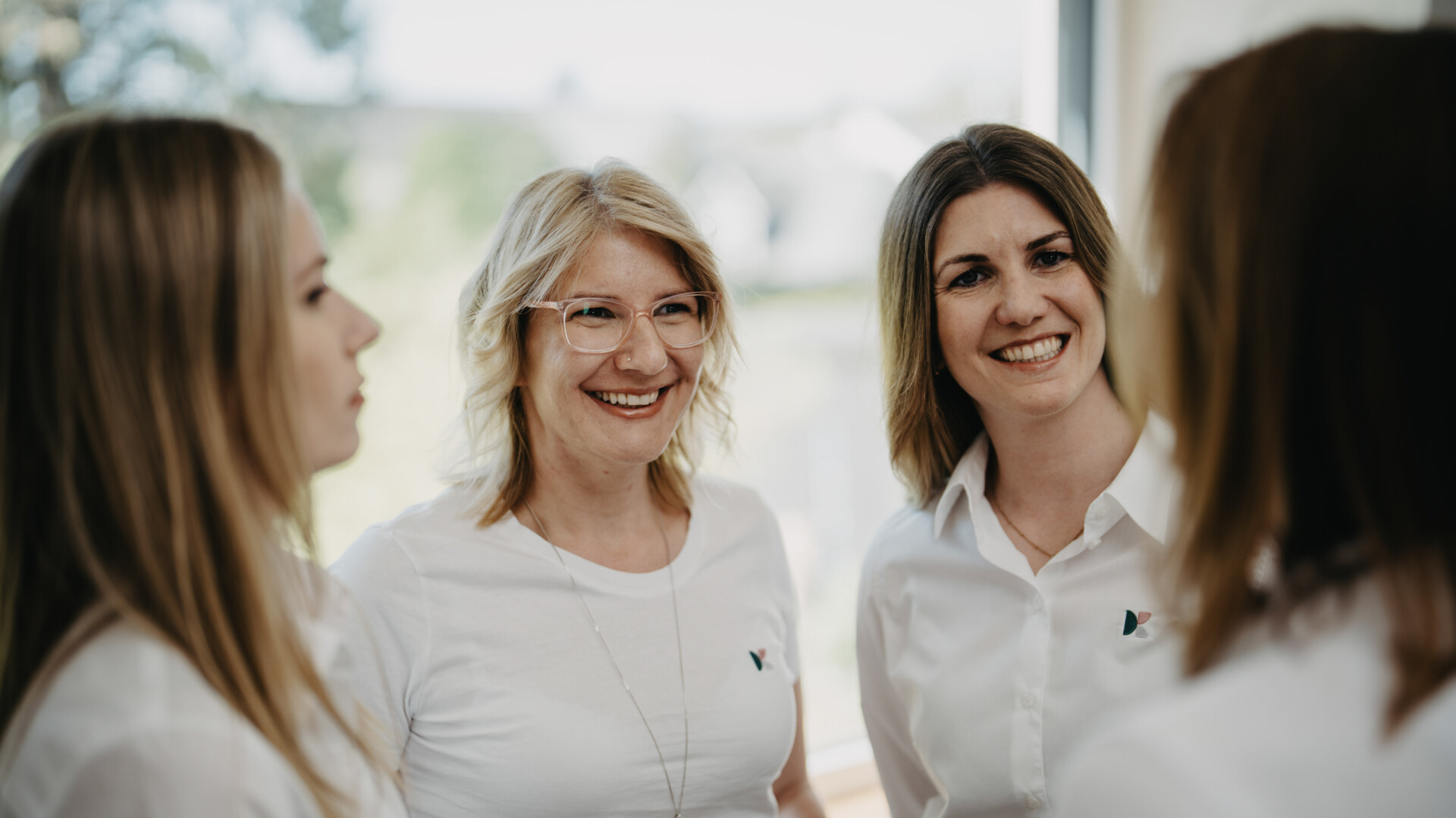 Vier Frauen in wei&szlig;en Oberteilen stehen in einer Gruppe zusammen und unterhalten sich l&auml;chelnd.