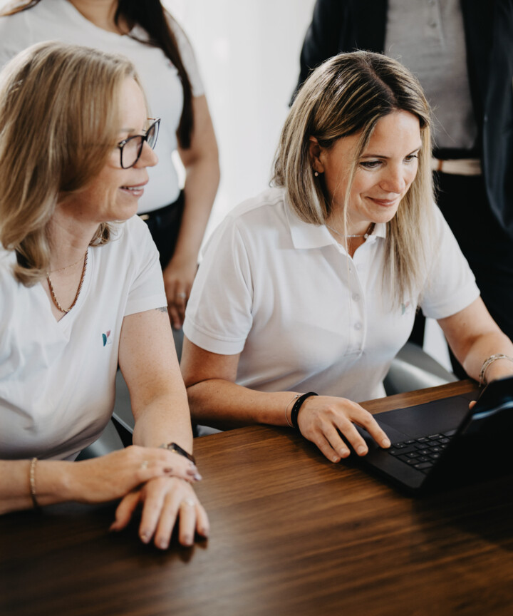 Vier Mitarbeiterinnen in wei&szlig;en Shirts und dunklem Blazer arbeiten l&auml;chelnd gemeinsam an einem Laptop.