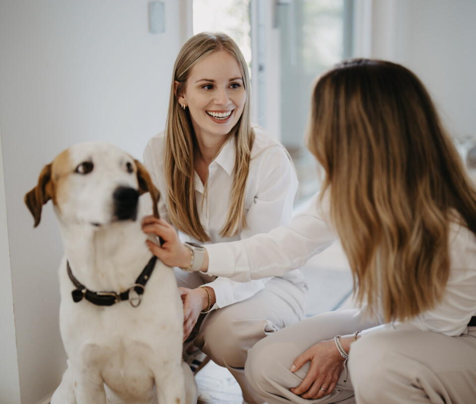 Anne G&ouml;ttlicher und eine Kollegin in wei&szlig;er Kleidung knien am Boden und streicheln einen hellen Hund mit braunen Flecken.