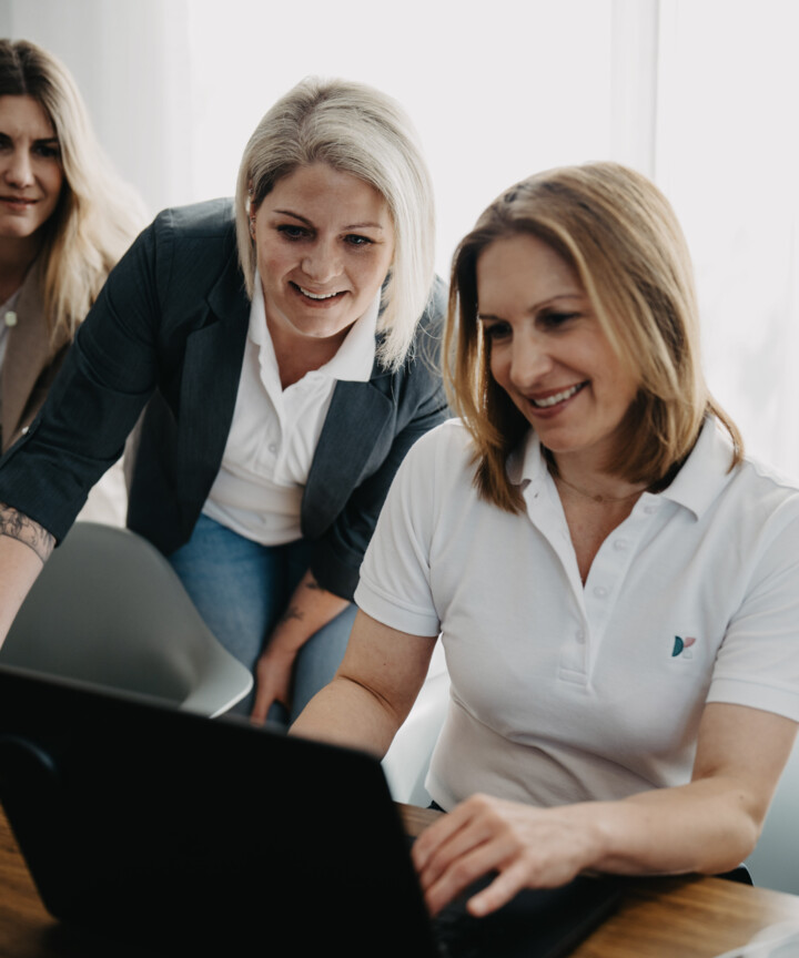 Christiane Berger arbeitet am Laptop, Charlene Antes im dunklen Blazer schaut ihr l&auml;chelnd &uuml;ber die Schulter.