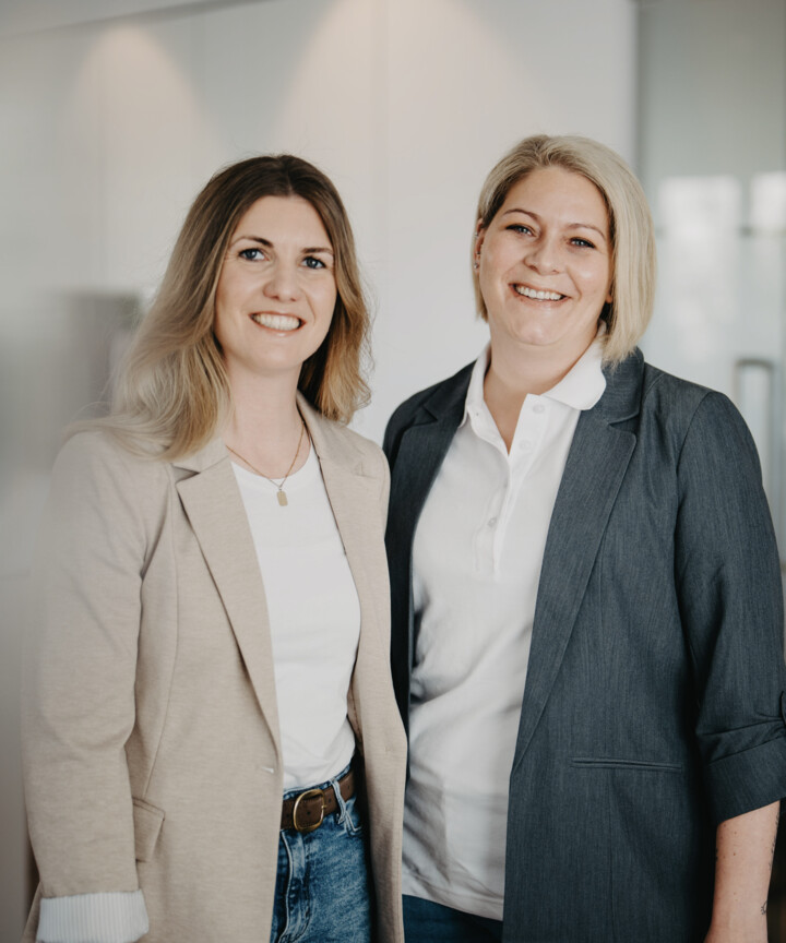 Zwei l&auml;chelnde Frauen im beigen und dunkelgrauen Blazer posieren nebeneinander f&uuml;r ein Portr&auml;tfoto.